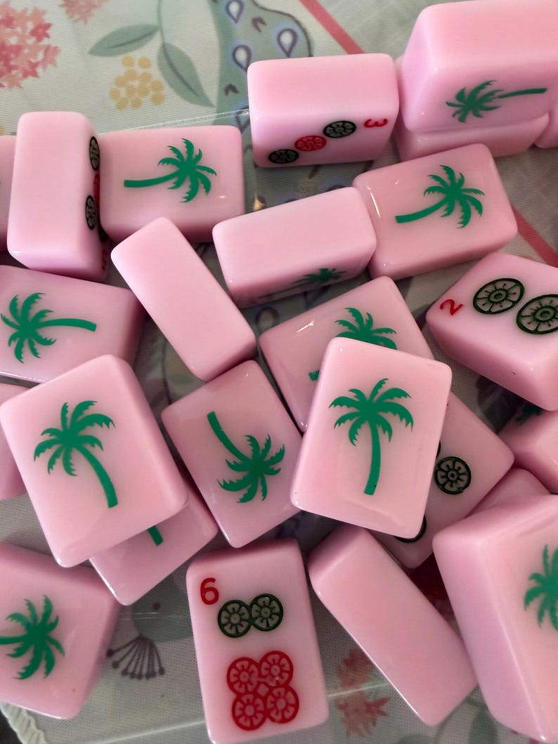 May include: Pink Mahjong tiles with green palm tree and other symbols. The tiles are scattered, showing various numbers and symbols. The tiles are a soft pink color, and the palm trees are green.