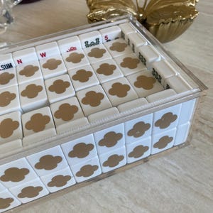 May include: A white tile game set with gold clover designs and other symbols, encased in a clear acrylic box. The game set is on a light-colored surface, with a gold butterfly decoration in the background.