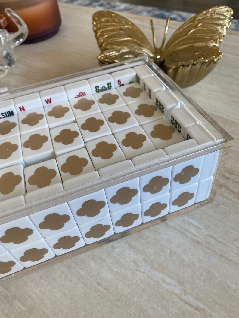 May include: A white tile game set with gold clover designs and other symbols, encased in a clear acrylic box. The game set is on a light-colored surface, with a gold butterfly decoration in the background.