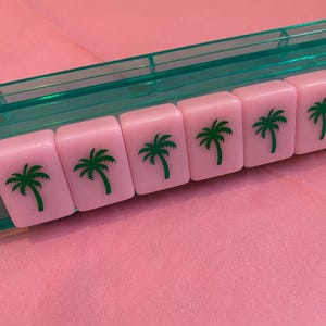 May include: Pink Mahjong tiles with green palm tree graphics are arranged in a row. The tiles are in a clear green plastic tray, set against a pink background. The image showcases a close-up of the tiles.