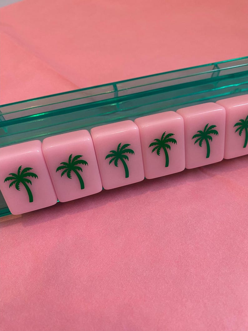 May include: Pink Mahjong tiles with green palm tree graphics are arranged in a row. The tiles are in a clear green plastic tray, set against a pink background. The image showcases a close-up of the tiles.