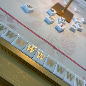 May include: A close-up of a Mahjong game in progress. The tiles are white with gold lettering and symbols. The game board has a floral design with pink and gold accents. A gold stand holds a gold square.