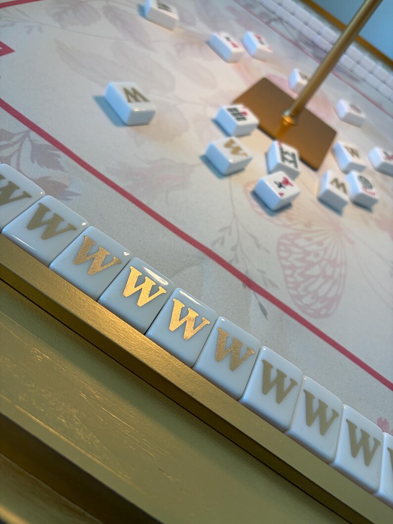 May include: A close-up of a Mahjong game in progress. The tiles are white with gold lettering and symbols. The game board has a floral design with pink and gold accents. A gold stand holds a gold square.