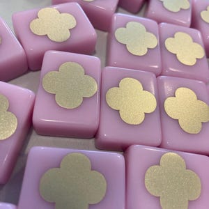 May include: A collection of pink, rectangular tiles with a gold four-leaf clover design on each. The tiles are arranged closely together, creating a pattern. The image is well-lit, highlighting the glossy surface of the tiles.