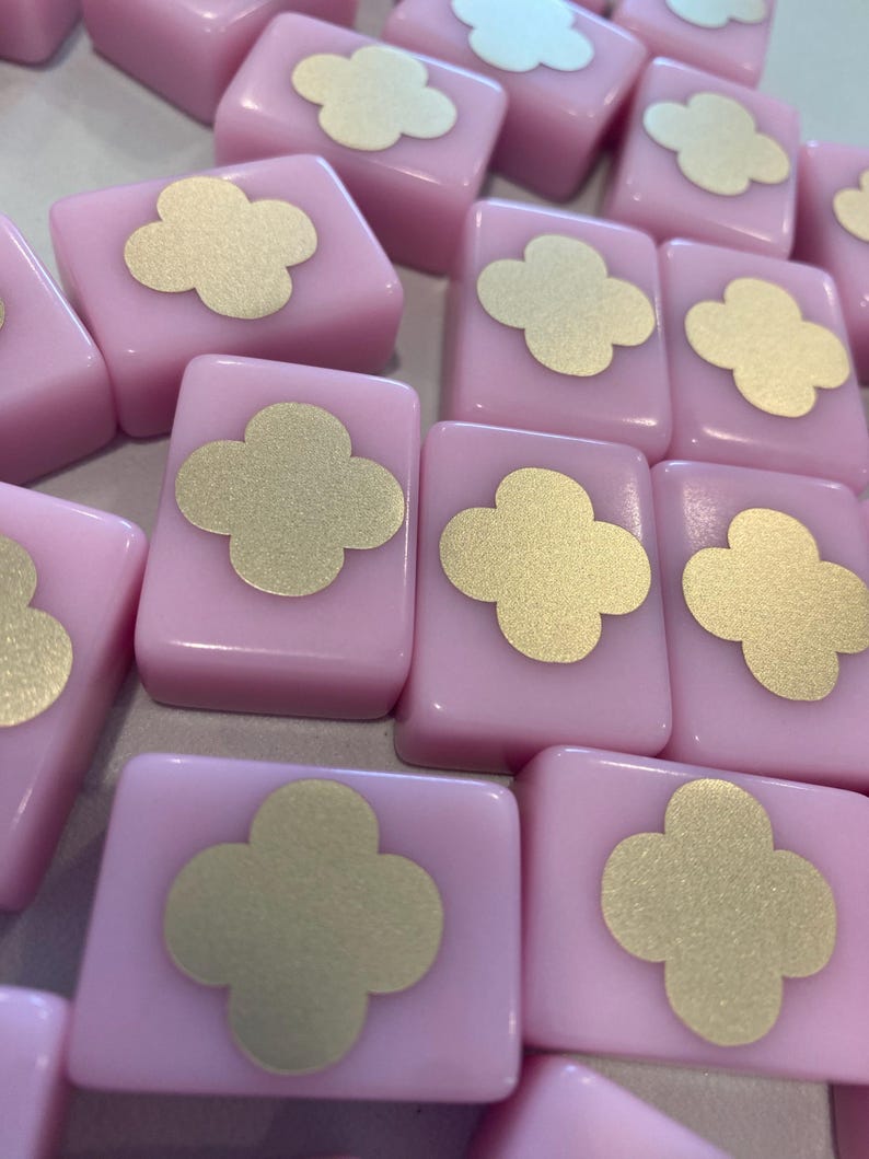 May include: A collection of pink, rectangular tiles with a gold four-leaf clover design on each. The tiles are arranged closely together, creating a pattern. The image is well-lit, highlighting the glossy surface of the tiles.