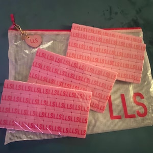 May include: Pink rectangular tiles with the letters "LLS" repeated across their surfaces are displayed. A clear mesh bag with the letters "LLS" printed on it is visible, along with a small pink heart-shaped charm with "LLS" on it.