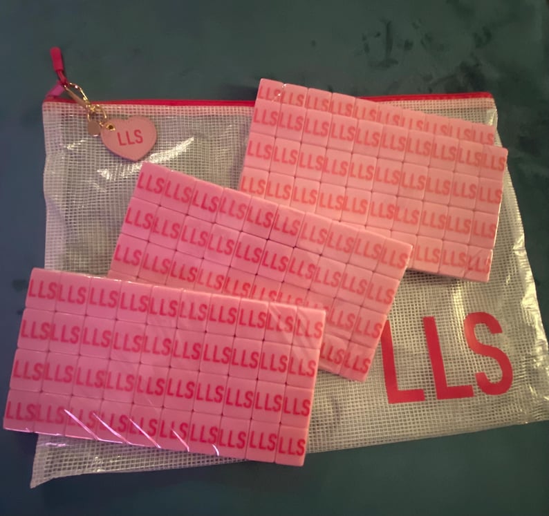 May include: Pink rectangular tiles with the letters "LLS" repeated across their surfaces are displayed. A clear mesh bag with the letters "LLS" printed on it is visible, along with a small pink heart-shaped charm with "LLS" on it.