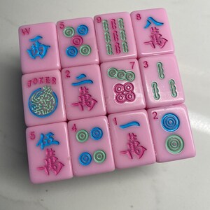 Pu&ograve; includere: Tessere rosa di Mahjong con simboli e numeri blu, verdi e rossi. Le tessere mostrano vari caratteri, numeri e simboli, inclusa la parola "Joker". Le tessere sono disposte in modo sparso.