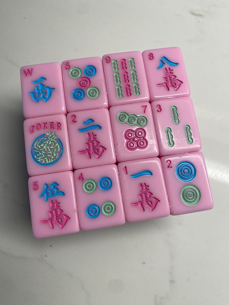 Pu&ograve; includere: Tessere rosa di Mahjong con simboli e numeri blu, verdi e rossi. Le tessere mostrano vari caratteri, numeri e simboli, inclusa la parola "Joker". Le tessere sono disposte in modo sparso.