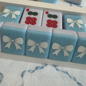 May include: A close-up of a set of Mahjong tiles. The tiles are light blue with white bow designs and white tops. Some tiles have red and green flower designs. The tiles are arranged in a white tray.