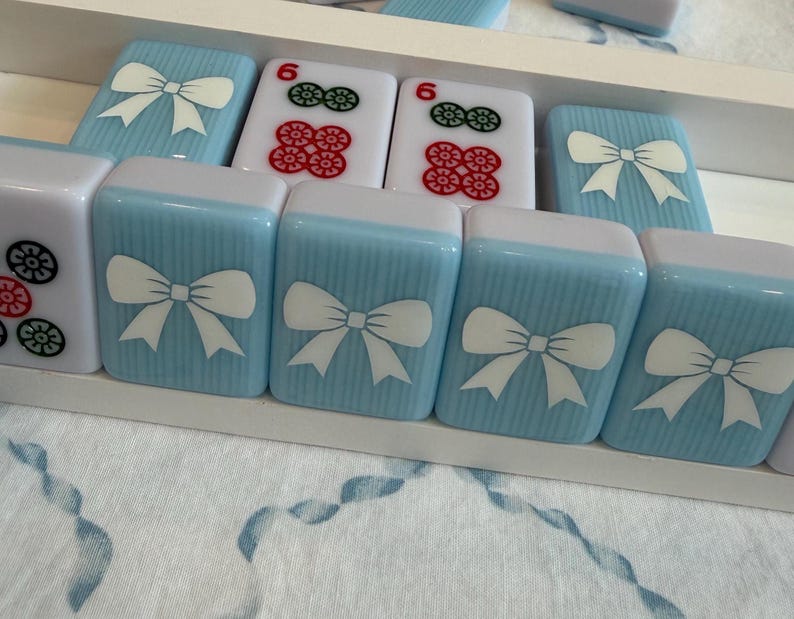 May include: A close-up of a set of Mahjong tiles. The tiles are light blue with white bow designs and white tops. Some tiles have red and green flower designs. The tiles are arranged in a white tray.