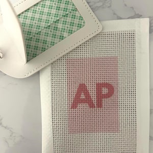 May include: A white luggage tag with a clear window and a green and white patterned insert is next to a white canvas with the letters "AP" in red. The canvas has a pink square behind the letters.