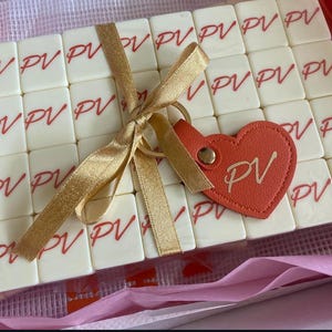 May include: A collection of off-white tiles, each printed with the letters "PV" in red, is tied with a gold ribbon. A heart-shaped, orange leather keychain with "PV" is attached. The tiles are arranged in a clear box, suggesting a personalized gift.