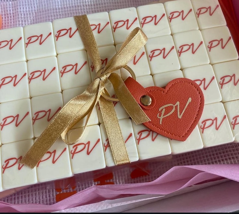 May include: A collection of off-white tiles, each printed with the letters "PV" in red, is tied with a gold ribbon. A heart-shaped, orange leather keychain with "PV" is attached. The tiles are arranged in a clear box, suggesting a personalized gift.
