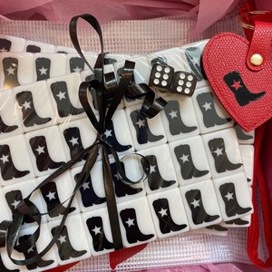 May include: A collection of white tiles featuring black cowboy boot and star motifs, secured with a black ribbon. Includes two dice and a red heart-shaped keyring with a cowboy boot design, presented in a pink box.