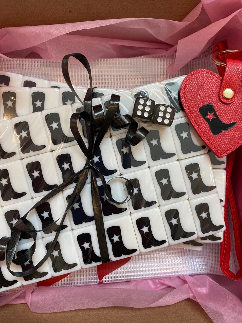 May include: A collection of white tiles featuring black cowboy boot and star motifs, secured with a black ribbon. Includes two dice and a red heart-shaped keyring with a cowboy boot design, presented in a pink box.