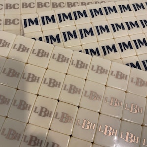 May include: Off-white rectangular tiles with printed initials. Some tiles display "JM" in navy blue, others show "LBH" in a metallic hue, and some have "BC" in a gray color. The tiles are arranged in multiple stacks.
