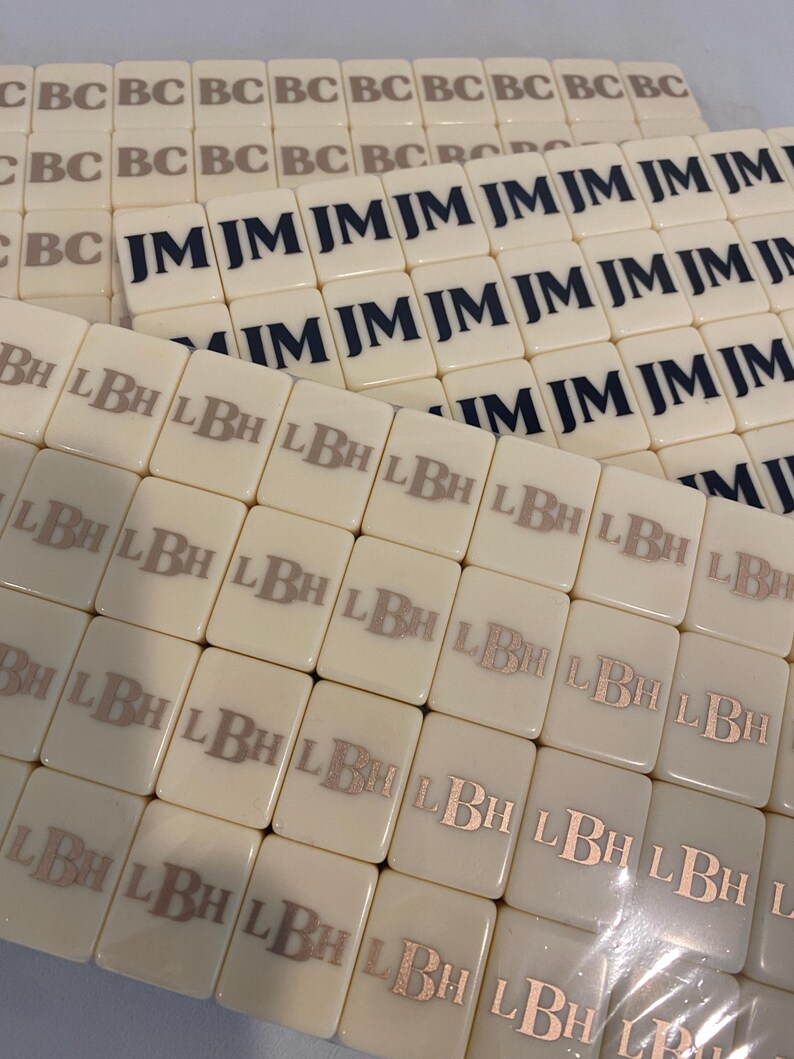 May include: Off-white rectangular tiles with printed initials. Some tiles display "JM" in navy blue, others show "LBH" in a metallic hue, and some have "BC" in a gray color. The tiles are arranged in multiple stacks.