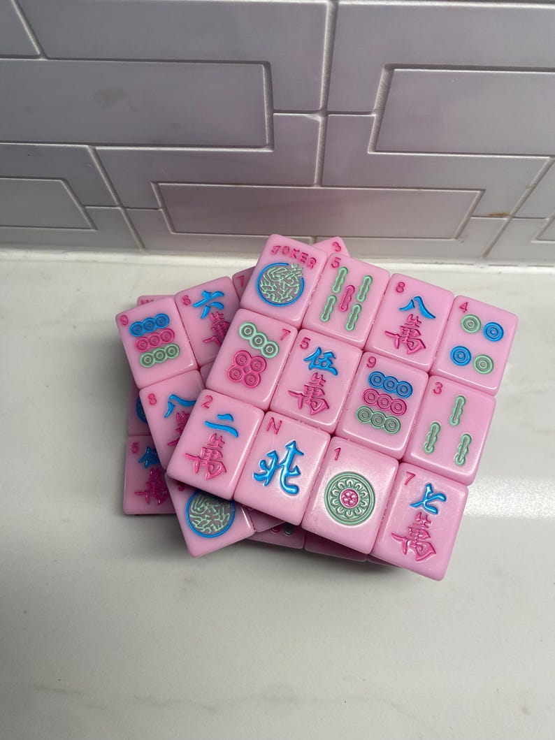 Pu&ograve; includere: Tessere rosa di Mahjong con simboli e caratteri blu, verdi e rossi. Le tessere sono impilate in modo casuale. Alcune tessere hanno la parola "Joker".