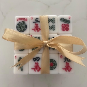 Pu&ograve; includere: Un set di tessere Mahjong bianche con simboli rossi, verdi e neri, legato con un nastro dorato. Le tessere includono la parola "JOKER" e numeri. Un set regalo decorativo per gli appassionati di giochi.