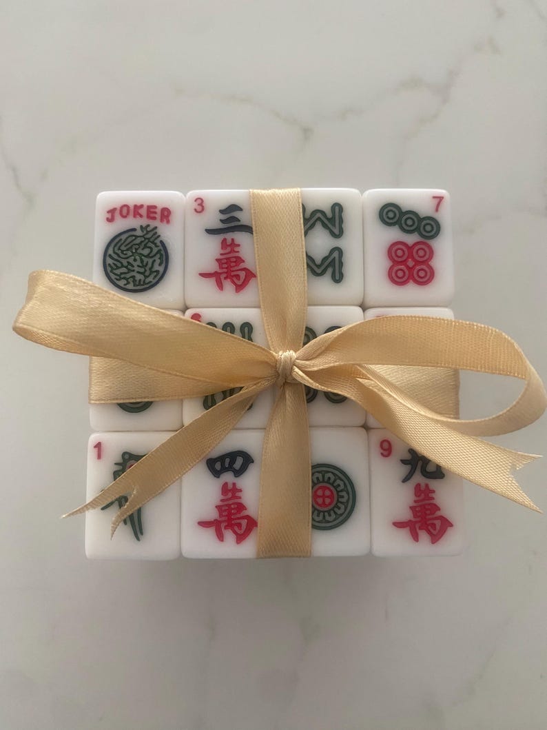 Pu&ograve; includere: Un set di tessere Mahjong bianche con simboli rossi, verdi e neri, legato con un nastro dorato. Le tessere includono la parola "JOKER" e numeri. Un set regalo decorativo per gli appassionati di giochi.