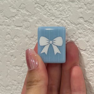 May include: A light blue, rectangular tile with a white bow design. The tile has vertical stripes and is held in a hand. The bow is centered on the tile. The background is a textured white wall.