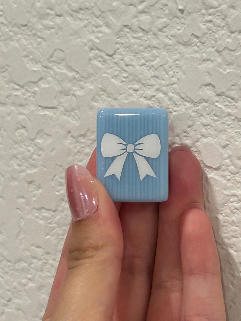 May include: A light blue, rectangular tile with a white bow design. The tile has vertical stripes and is held in a hand. The bow is centered on the tile. The background is a textured white wall.