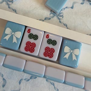 May include: A set of light blue and white Mahjong tiles. Some tiles have a white bow design, while others display the number 6 with red and green symbols. The tiles are arranged in a white tray.