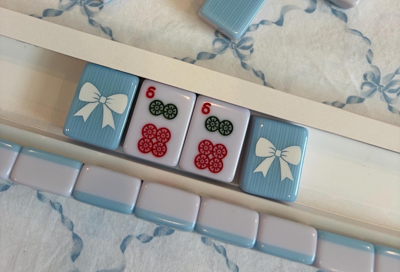 May include: A set of light blue and white Mahjong tiles. Some tiles have a white bow design, while others display the number 6 with red and green symbols. The tiles are arranged in a white tray.
