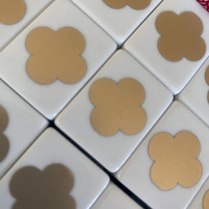May include: Close-up of white Mahjong tiles with gold-colored four-leaf clover symbols. The tiles are arranged in a diagonal pattern, showcasing the details of the game pieces. The gold clovers are centered on each tile.