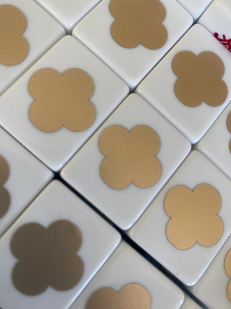 May include: Close-up of white Mahjong tiles with gold-colored four-leaf clover symbols. The tiles are arranged in a diagonal pattern, showcasing the details of the game pieces. The gold clovers are centered on each tile.