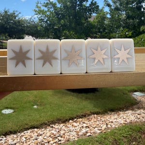 May include: Five rectangular off-white tiles arranged in a row on a gold-colored ledge. Each tile features a centered, metallic gold eight-pointed star design. The tiles have a smooth, glossy finish and are positioned horizontally against a background.