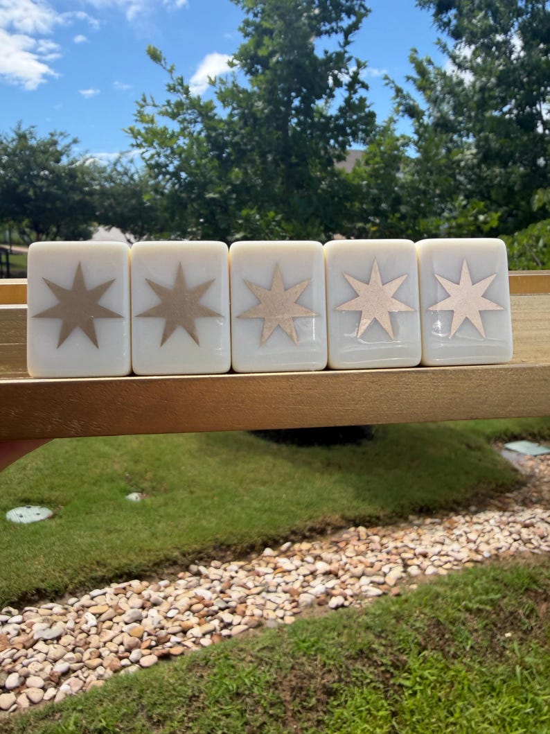 May include: Five rectangular off-white tiles arranged in a row on a gold-colored ledge. Each tile features a centered, metallic gold eight-pointed star design. The tiles have a smooth, glossy finish and are positioned horizontally against a background.