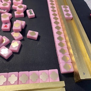 May include: A pink Mahjong set with gold accents. The tiles are pink with gold floral designs and Chinese characters. The set includes gold-colored racks and a black playing surface.