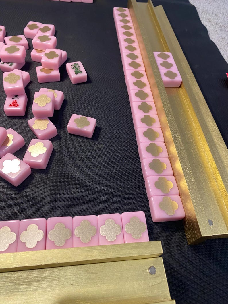 May include: A pink Mahjong set with gold accents. The tiles are pink with gold floral designs and Chinese characters. The set includes gold-colored racks and a black playing surface.