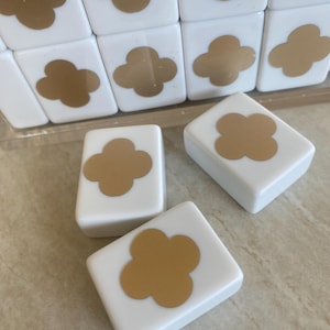 May include: White square tiles with a gold clover design. The tiles are arranged in a grid, with some tiles scattered in front. The clover design is centered on each tile. The tiles appear to be made of ceramic or a similar material.