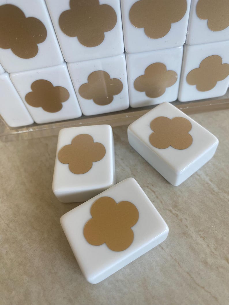 May include: White square tiles with a gold clover design. The tiles are arranged in a grid, with some tiles scattered in front. The clover design is centered on each tile. The tiles appear to be made of ceramic or a similar material.