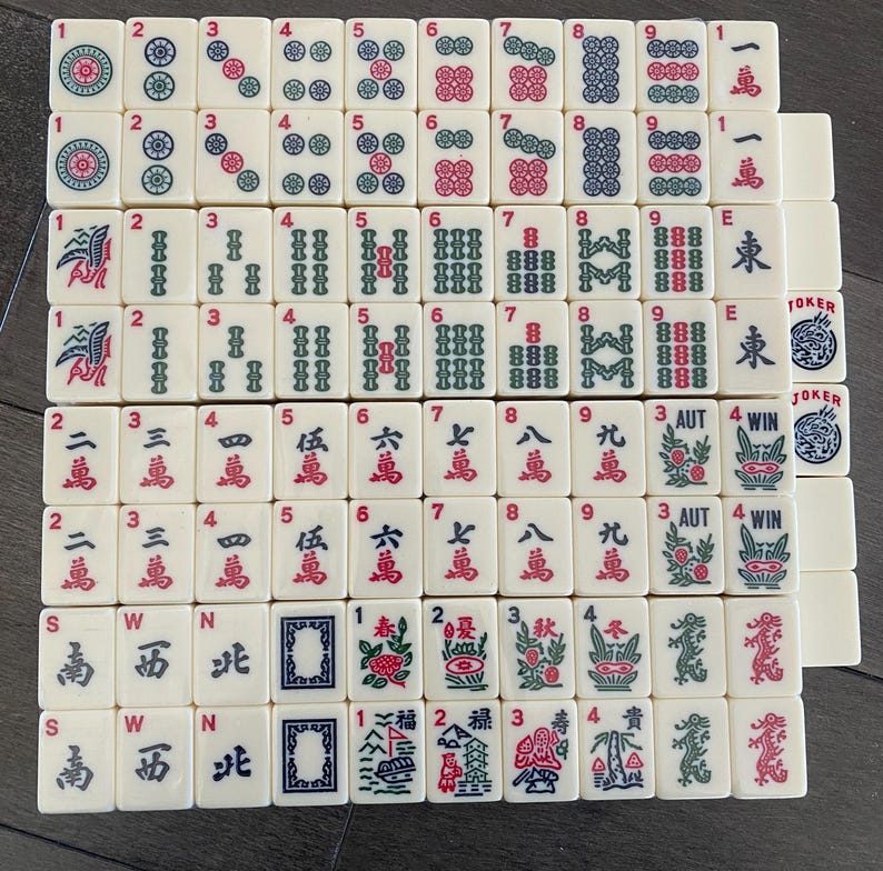 May include: An assortment of ivory-coloured Mahjong tiles, each featuring distinct symbols, numbers, and Chinese characters in red, green, and blue. The tiles are arranged in a grid, displaying the variety of designs and suits.
