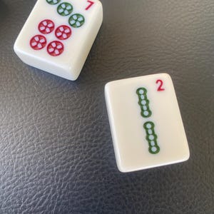 May include: Two white Mahjong tiles. One tile shows the number 7 in red, with a pattern of green and red flower-like symbols. The other tile shows the number 2 in red, with a pattern of green symbols.