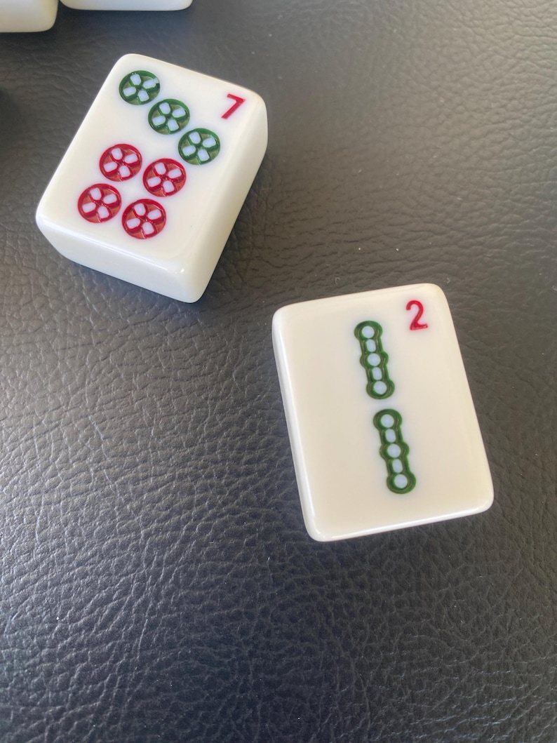May include: Two white Mahjong tiles. One tile shows the number 7 in red, with a pattern of green and red flower-like symbols. The other tile shows the number 2 in red, with a pattern of green symbols.