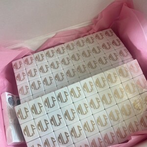 May include: White tiles with a gold monogram, arranged in a box with pink tissue paper. The tiles are square and feature the letters "MLF". A pack of cards is visible on the left side of the box.