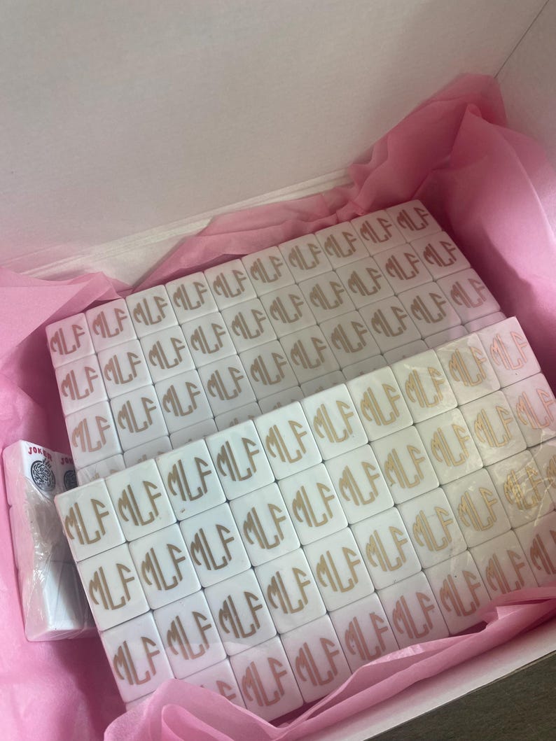 May include: White tiles with a gold monogram, arranged in a box with pink tissue paper. The tiles are square and feature the letters "MLF". A pack of cards is visible on the left side of the box.