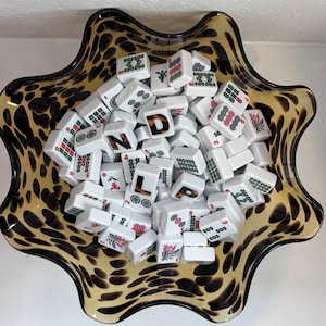 May include: A scalloped glass bowl with a brown and black leopard print pattern filled with a collection of white rectangular mahjong tiles. The tiles feature various green and red symbols, numbers, and the letters D, N, L, and P in a brown font.