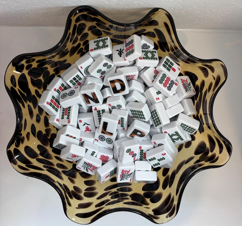 May include: A scalloped glass bowl with a brown and black leopard print pattern filled with a collection of white rectangular mahjong tiles. The tiles feature various green and red symbols, numbers, and the letters D, N, L, and P in a brown font.