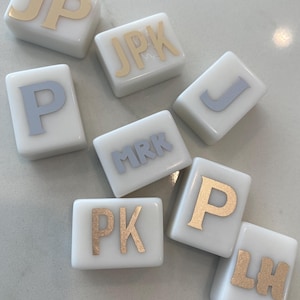 May include: White square tiles with gold and silver lettering spelling out initials. The letters include JP, JPK, P, MRK, J, PK, P, and LH. The tiles are arranged on a light-colored surface.