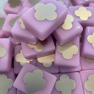 May include: A pile of light pink square tiles with a gold clover design in the center. The tiles are stacked together, creating a textured surface. The gold clover design adds a touch of elegance to the pink tiles.