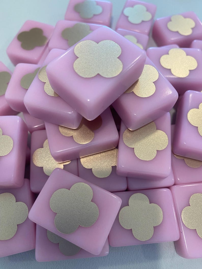 May include: A pile of light pink square tiles with a gold clover design in the center. The tiles are stacked together, creating a textured surface. The gold clover design adds a touch of elegance to the pink tiles.