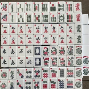 May include: A collection of white Mahjong tiles, featuring red and green symbols and numbers. The tiles display various suits, including characters, bamboos, and circles, along with directional winds and Jokers. A classic game.