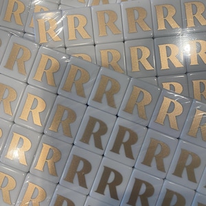 May include: A collection of white square tiles, each featuring a gold letter "R". The tiles are arranged in a grid pattern, suggesting a game or craft supply. The gold lettering contrasts with the white background.