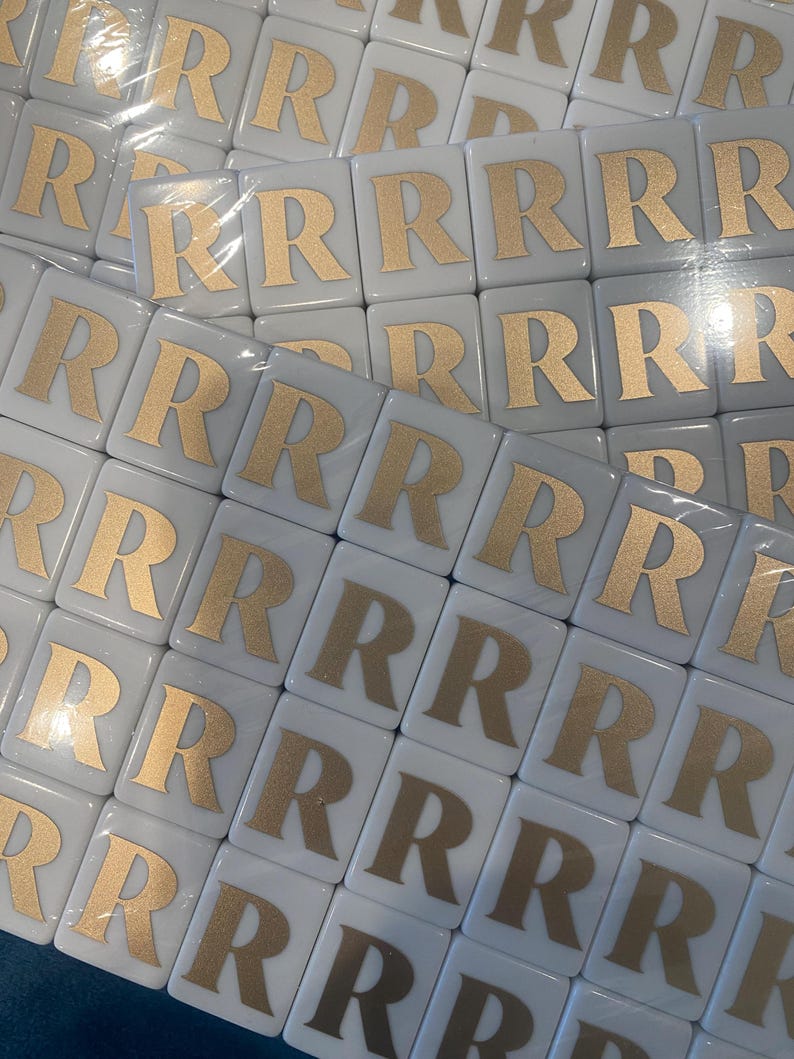 May include: A collection of white square tiles, each featuring a gold letter "R". The tiles are arranged in a grid pattern, suggesting a game or craft supply. The gold lettering contrasts with the white background.